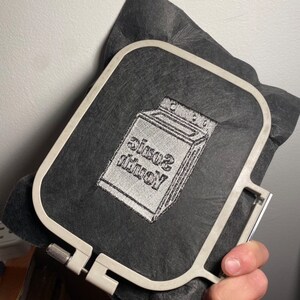 May include: A gray embroidery hoop holding a black fabric with a white embroidered design. The design features a washing machine with the words "Sonic Youth" in a stylized font. The hoop is held in a hand.