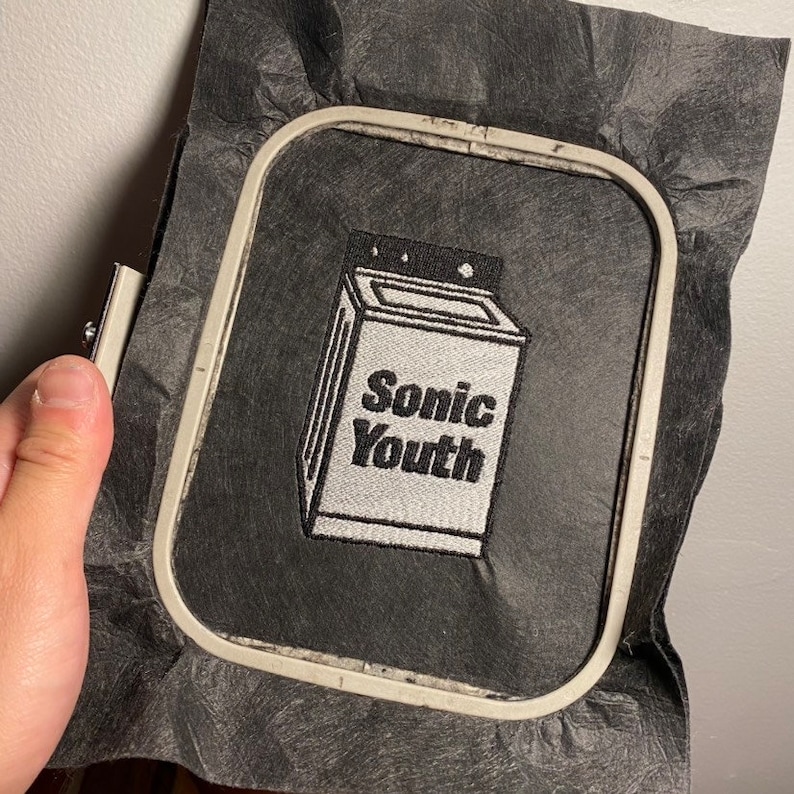May include: Embroidered patch featuring a washing machine design with the text "Sonic Youth" in black and white. The patch is set within an embroidery hoop, on a black fabric background.
