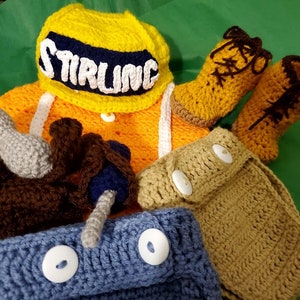 May include: A collection of handmade crochet items, including a yellow hat with "STIRLING" in white letters, an orange vest, brown boots, and blue and tan accessories. The items are arranged on a green surface.