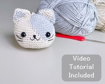 Może przedstawiać: Pluszowa zabawka amigurumi w kształcie kota, wykonana z białej i szarej włóczki. Zabawka ma czarne oczy i czarny nos. Na zdjęciu widoczne są również motki włóczki i szydełko. Tekst "Samouczek wideo w zestawie" znajduje się na szarym tle.