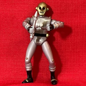 May include: A silver action figure of a robot-like character with a gold-colored head and accents. The figure is holding a silver and white weapon. It has black boots with gold details and is posed against a red background.