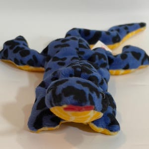 May include: A plush toy frog in a lying position. The frog is blue with black spots and yellow accents on the feet and belly. It has a yellow mouth and red lips. The toy is soft and cuddly.