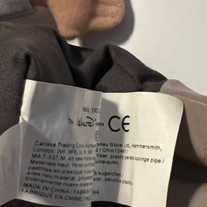 May include: A close-up of a brown and gray plush toy with a white tag. The tag has the text "The Walt Disney Company" and "CE" in a circle. The tag also has the text "Canasa Trading Corp. Burton sney Store Ltd., Hammersmith, London W6 9PE, UK. ME 2/Ohio 12441/ MA T-537-M, All new material, fiber, plastic pellets sponge pipe/ Materiels neut, fibres synthetiques, pastilles en plastique,  kugeichen/ Alles neu material, polyester, pelota plastico Enteramente nuevo material, FABRIQUE EN CHINE/HE"