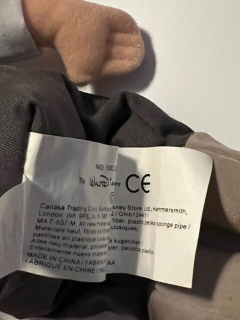 May include: A close-up of a brown and gray plush toy with a white tag. The tag has the text "The Walt Disney Company" and "CE" in a circle. The tag also has the text "Canasa Trading Corp. Burton sney Store Ltd., Hammersmith, London W6 9PE, UK. ME 2/Ohio 12441/ MA T-537-M, All new material, fiber, plastic pellets sponge pipe/ Materiels neut, fibres synthetiques, pastilles en plastique,  kugeichen/ Alles neu material, polyester, pelota plastico Enteramente nuevo material, FABRIQUE EN CHINE/HE"