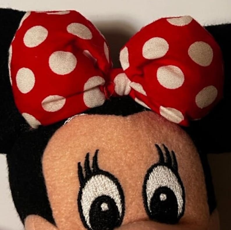 May include: A plush toy of a cartoon mouse with black ears, a red bow with white polka dots, and embroidered eyes. The mouse has a pink face and black eyelashes.