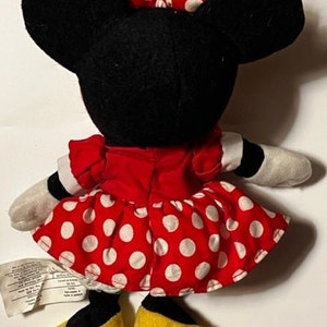 May include: A red and white polka dot dress with a black bow on top of the head. The plush toy is wearing yellow shoes.