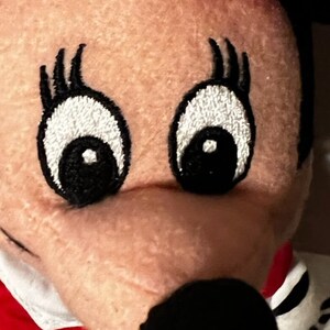 May include: Close-up of a plush toy's face with embroidered eyes and eyelashes. The toy has a red and white polka dot bow and a black nose.