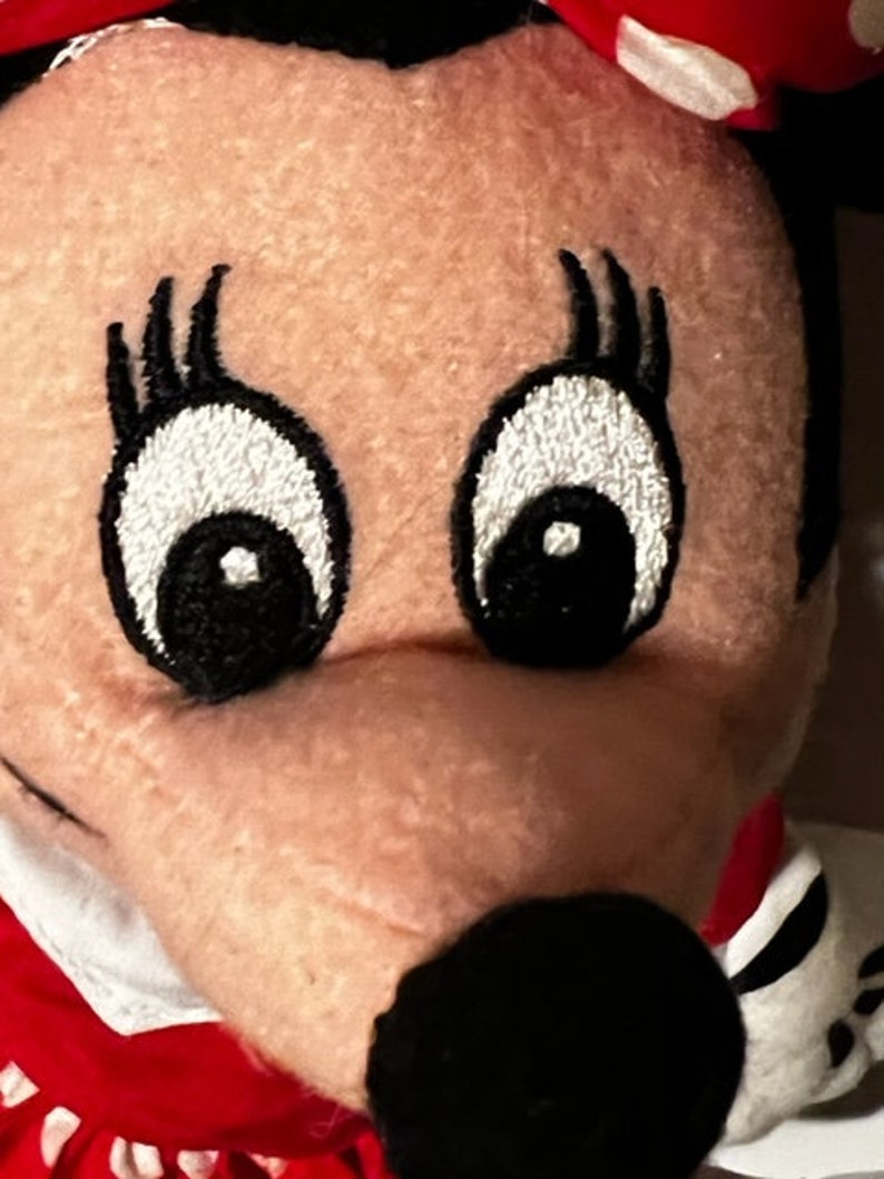 May include: Close-up of a plush toy's face with embroidered eyes and eyelashes. The toy has a red and white polka dot bow and a black nose.