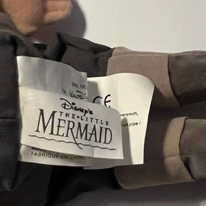 May include: A plush toy of the character Ursula from Disney's The Little Mermaid. The toy is brown and gray with a white tag that reads "Disney's The Little Mermaid" and "Fabrique en Chine".