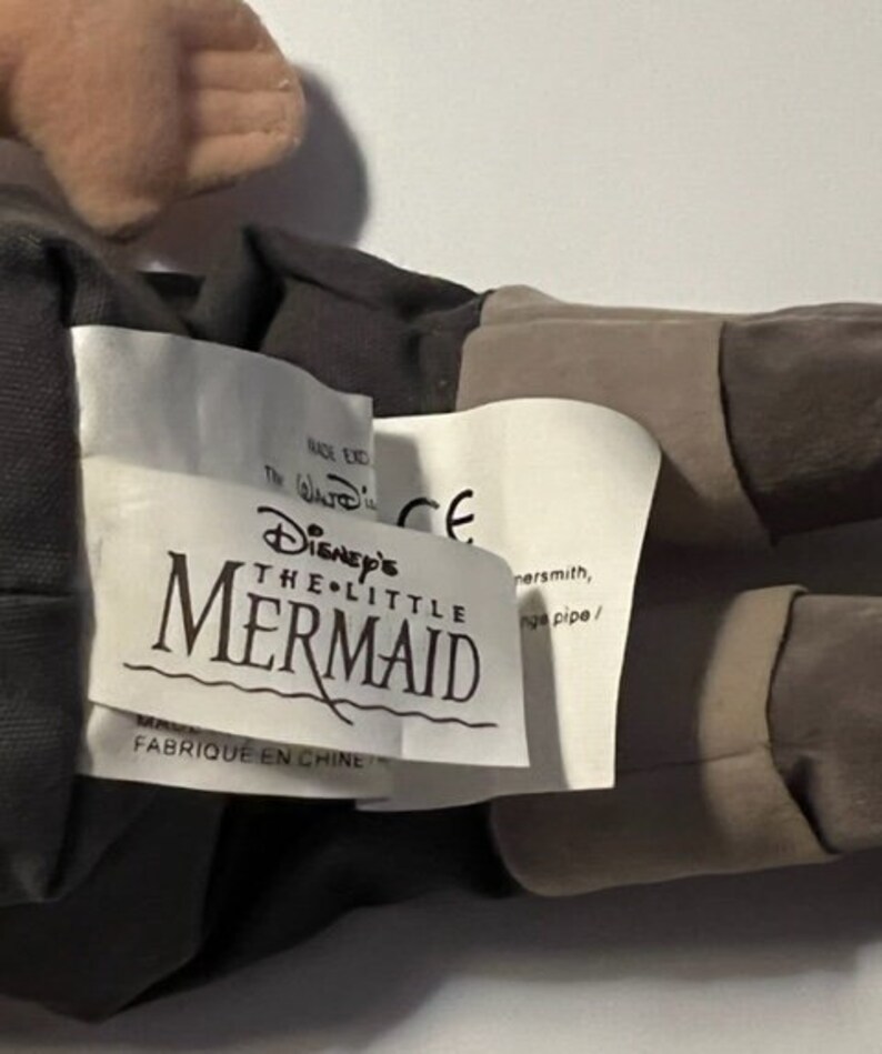 May include: A plush toy of the character Ursula from Disney's The Little Mermaid. The toy is brown and gray with a white tag that reads "Disney's The Little Mermaid" and "Fabrique en Chine".