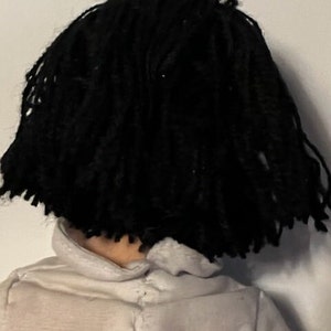 May include: A doll with black yarn hair styled in dreadlocks. The doll is wearing a white shirt.