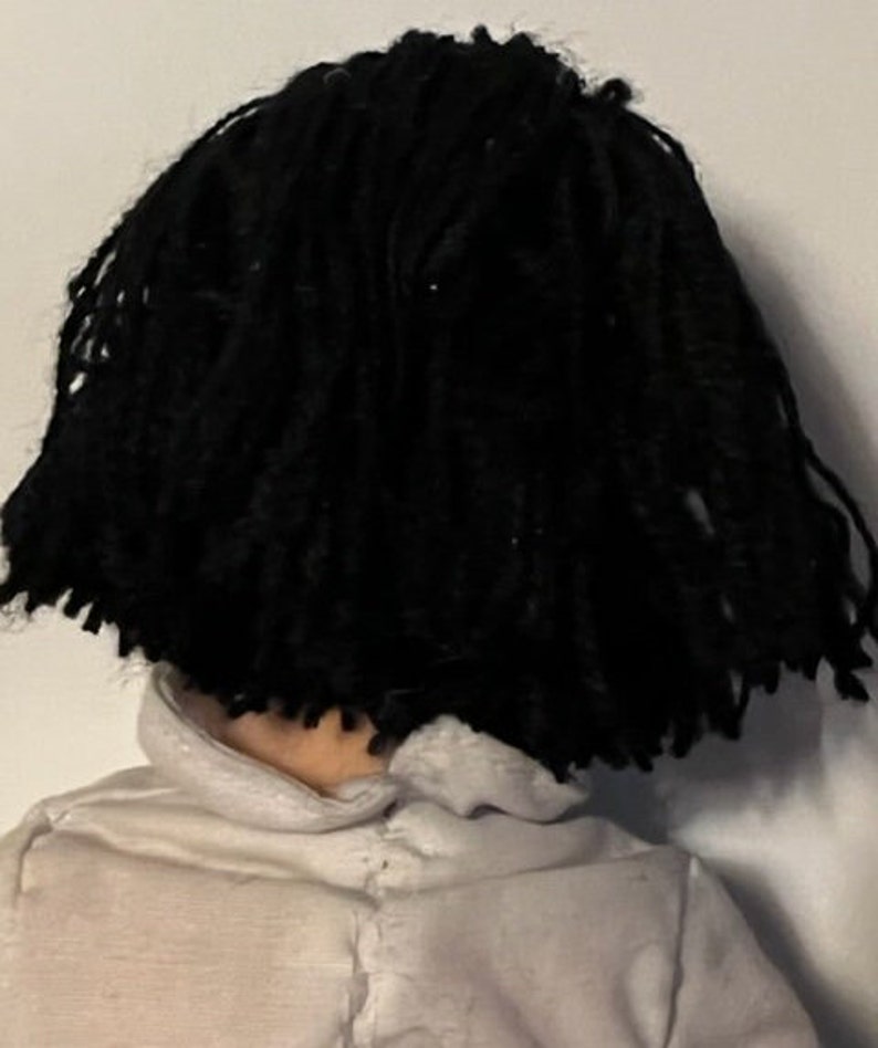 May include: A doll with black yarn hair styled in dreadlocks. The doll is wearing a white shirt.