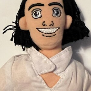 May include: A plush toy of a man with black hair, wearing a white shirt with a collar. He has a smiling face with embroidered features.