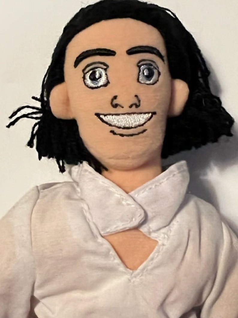 May include: A plush toy of a man with black hair, wearing a white shirt with a collar. He has a smiling face with embroidered features.