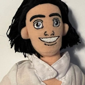 May include: A plush doll with black hair, a white shirt, and a smiling face. The doll has embroidered eyes and a mouth.