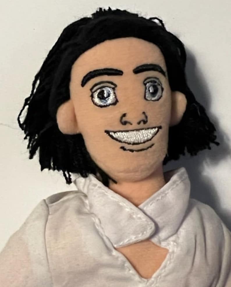 May include: A plush doll with black hair, a white shirt, and a smiling face. The doll has embroidered eyes and a mouth.