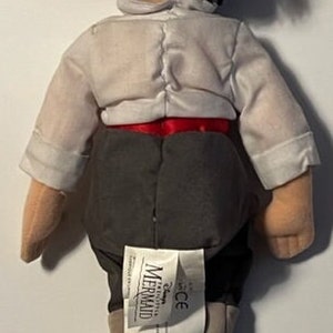 May include: A plush toy of Prince Eric from Disney's The Little Mermaid. He is wearing a white shirt, red sash, gray pants, and brown boots. The toy has a Disney The Little Mermaid tag attached to the back.