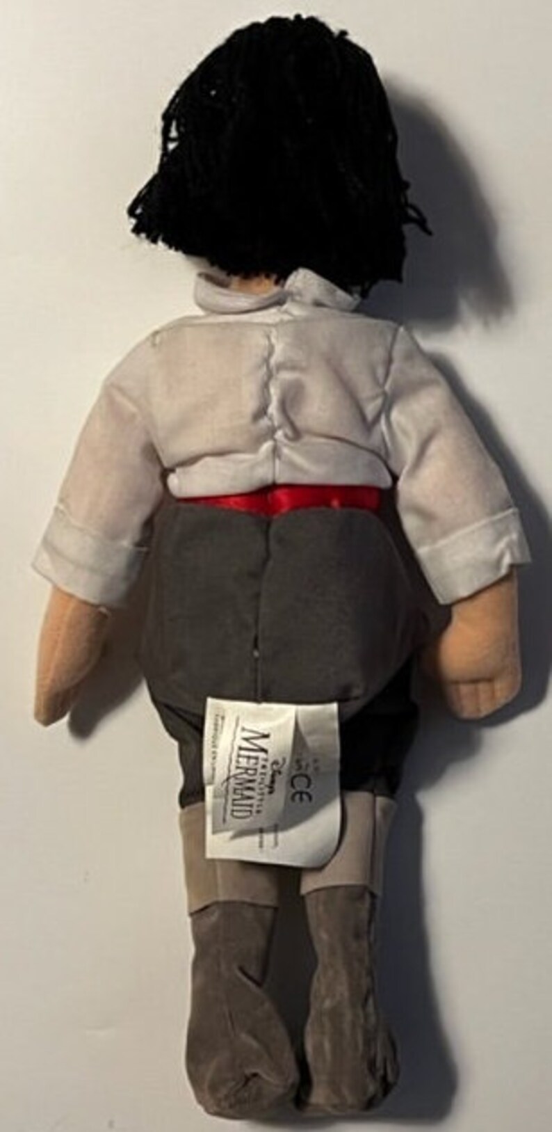 May include: A plush toy of Prince Eric from Disney's The Little Mermaid. He is wearing a white shirt, red sash, gray pants, and brown boots. The toy has a Disney The Little Mermaid tag attached to the back.