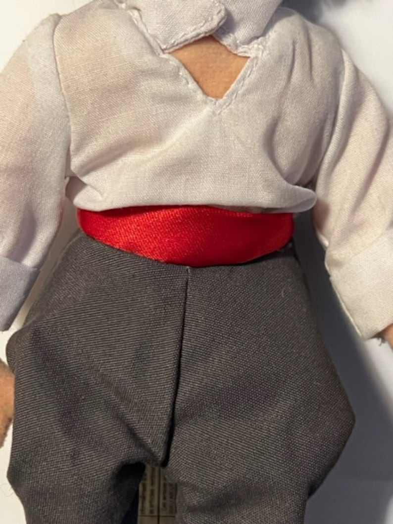 May include: A white shirt with a red sash around the waist and black pants. The shirt has a V-neck and a small opening at the neckline.