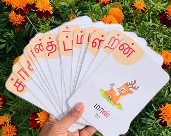 May include: A set of white flashcards with Tamil letters printed in red. The cards are fanned out in a hand, with the top card showing a cartoon illustration of a deer and the Tamil word for deer, "மான்", which translates to "Maan".
