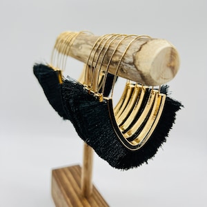 May include: A pair of gold hoop earrings with black fringe tassels hanging from a wooden earring stand.