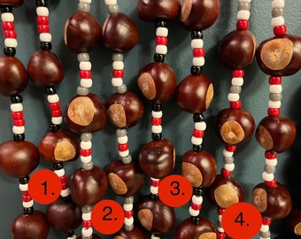May include: Close-up of a decorative hanging made of large, dark brown buckeye nuts. The nuts are strung together with alternating sections of red, white, gray, and black beads, creating a unique and natural aesthetic. The background is a muted teal color.