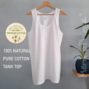 May include: White tank top made of 100% natural pure cotton. The tank top is hanging on a wooden hanger against a gray wall. The text "100% NATURAL", "BEST CHOICE", "PREMIUM COTTON", "100% NATURAL", "PURE COTTON", and "TANK TOP" is printed on the image.