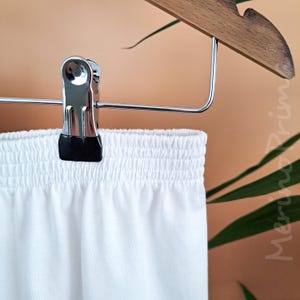 May include: White cotton shorts with an elastic waistband. The shorts are hanging on a wooden hanger with a silver clip.