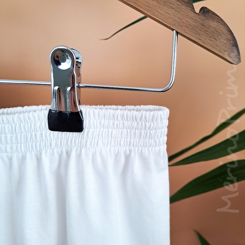 May include: White cotton shorts with an elastic waistband. The shorts are hanging on a wooden hanger with a silver clip.