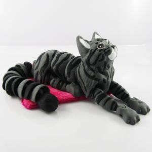May include: A grey and black tabby cat figurine with a curled tail, lying on a pink fabric mat. The cat has a white patch on its chest and is looking upwards.