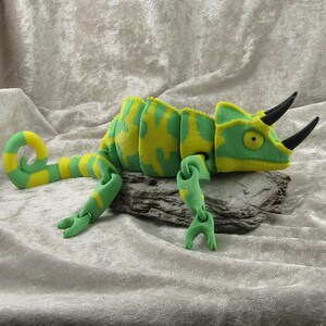 May include: A green and yellow plush chameleon toy with a curled tail and articulated limbs. The toy features a detailed design, including horns and a textured body, and is positioned on a piece of wood.