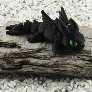 May include: A black 3D printed figurine of a dragon with green eyes. The dragon is lying on a piece of driftwood.
