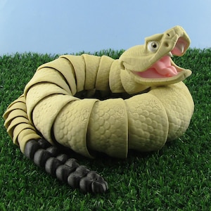 May include: A coiled, pale green snake ornament with an open mouth showing a pink interior. The snake's tail is black. The ornament is placed on artificial green grass against a light blue backdrop.