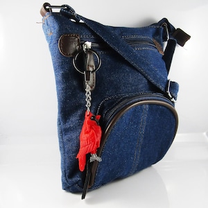 May include: A blue denim crossbody bag with a brown leather trim and a red cardinal keychain. The bag has a zippered pocket on the front and a flap closure.