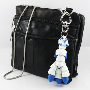 May include: A white and blue polymer clay charm shaped like a creature with a long tail and multiple legs. The charm is attached to a silver chain and a silver heart-shaped clasp. The charm is hanging from a black leather purse.