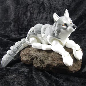 May include: A gray and white articulated wolf figurine with a segmented tail, lying on a piece of driftwood.