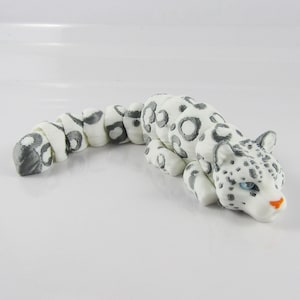 May include: A white and gray snow leopard figurine with a curled tail. The figurine is made of a smooth, white material with gray spots.