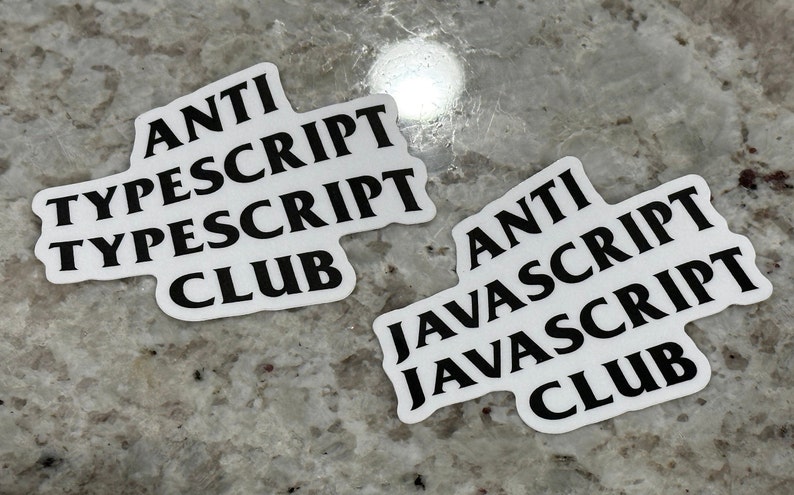 May include: Two white stickers with black text. The first sticker says "ANTI TYPESCRIPT TYPESCRIPT CLUB". The second sticker says "ANTI JAVASCRIPT JAVASCRIPT CLUB".