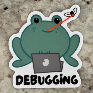 May include: A sticker featuring a cartoon frog with a laptop, a fly, and the word "DEBUGGING." The frog is green with black eyes and a smile. A red laser beam extends from the frog's nose to the fly. The laptop is gray with a white apple logo.