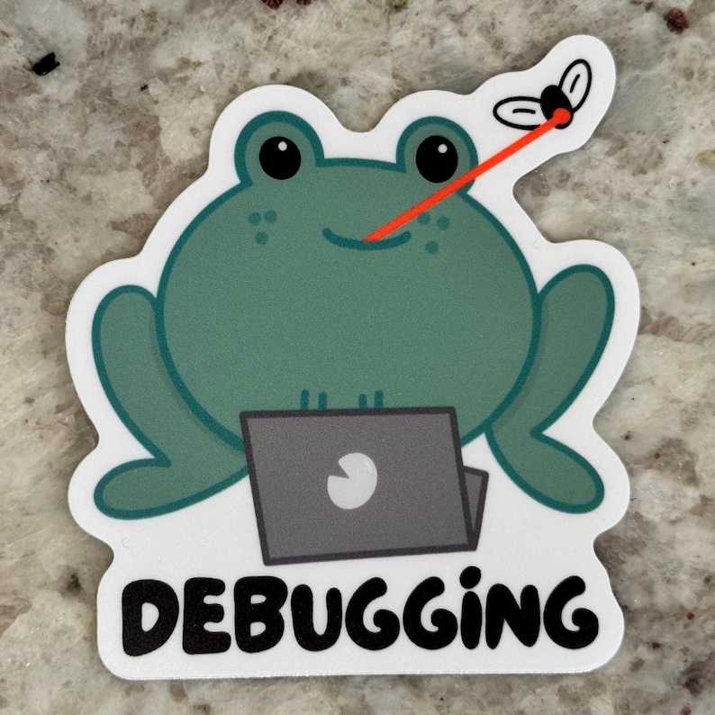 以下が含まれることがあります： ラップトップ、ハエ、そして「DEBUGGING」という言葉を持つ漫画のカエルのステッカー。カエルは緑色で、黒い目と笑顔をしています。赤いレーザービームがカエルの鼻からハエに伸びています。