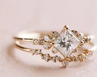May include: A gold ring with a large, square-cut diamond surrounded by smaller diamonds. The ring is set on a delicate band with a smaller band underneath.