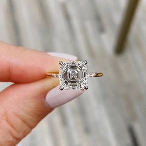 May include: A gold engagement ring featuring a square-shaped diamond. The diamond has a step-cut, also known as an emerald cut.
