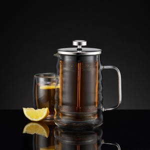 May include: A clear glass French press with a stainless steel lid and handle, filled with tea. A double-walled glass with tea and a lemon slice sits beside it. A lemon wedge rests in front.