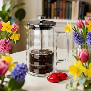 May include: A clear glass French press coffee maker with a stainless steel lid and plunger. The coffee maker is filled with dark coffee. Two red heart-shaped decorations are in front of the coffee maker. Colorful flowers surround the coffee maker.