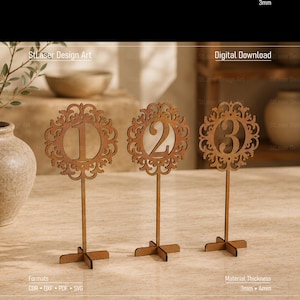 May include: Three brown laser-cut wooden table numbers labeled 1, 2, and 3. Each number is centered within a circular, ornate, filigree-style frame mounted on a thin vertical stem and a cross-shaped base. The text 3mm, Digital Download, Formats CDR, DXF, PDF, SVG, and Material Thickness 3mm, 4mm are visible.