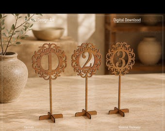 May include: Three brown laser-cut wooden table numbers labelled 1, 2, and 3. Each number is centred within a circular, ornate, filigree-style frame mounted on a thin vertical stem and a cross-shaped base. The text 3mm, Digital Download, Formats CDR, DXF, PDF, SVG, and Material Thickness 3mm, 4mm are visible.