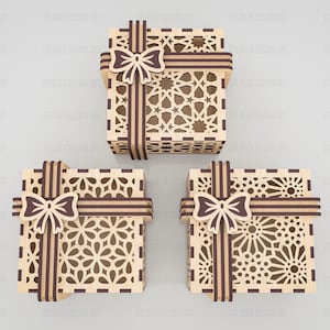 May include: Three square gift boxes made of light-colored wood with dark brown accents. Each box features intricate laser-cut geometric patterns on the lid and sides, topped with a decorative dark brown wooden bow and ribbon detail.