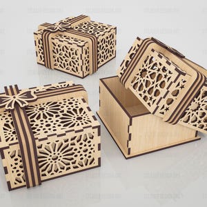 May include: Three square wooden gift boxes featuring intricate laser-cut geometric patterns on the lids and sides. Each box includes a decorative wooden ribbon and bow detail on top. One box is shown open with the lid resting beside the base.