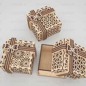 May include: Three square wooden gift boxes with intricate laser-cut geometric patterns on the lids and sides. Each box features a decorative wooden ribbon and bow design in a dark brown finish, with one box shown open to reveal a plain interior.