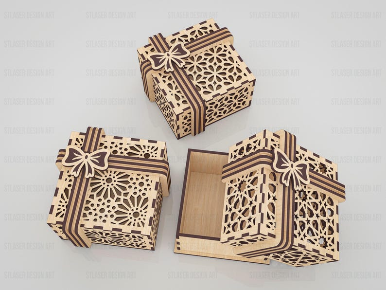 May include: Three square wooden gift boxes with intricate laser-cut geometric patterns on the lids and sides. Each box features a decorative wooden ribbon and bow design in a dark brown finish, with one box shown open to reveal a plain interior.